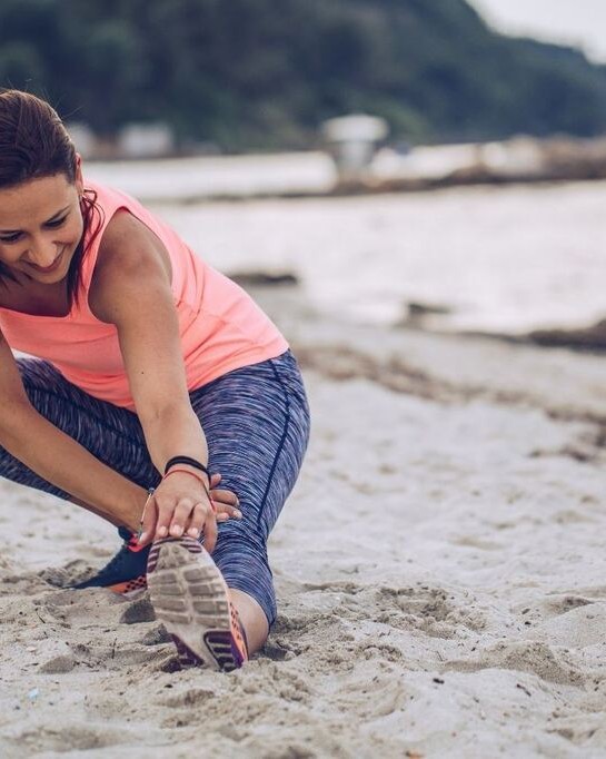 Mulher fazendo alongamento em treino na praia, com roupa leve e confortável para movimentos ao ar livre.