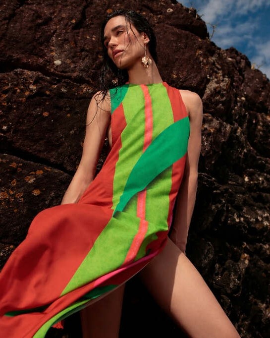 Mulher usando vestido leve estampado De Chelles encostada em pedra e com céu azul ao fundo, representando como usar saída de praia