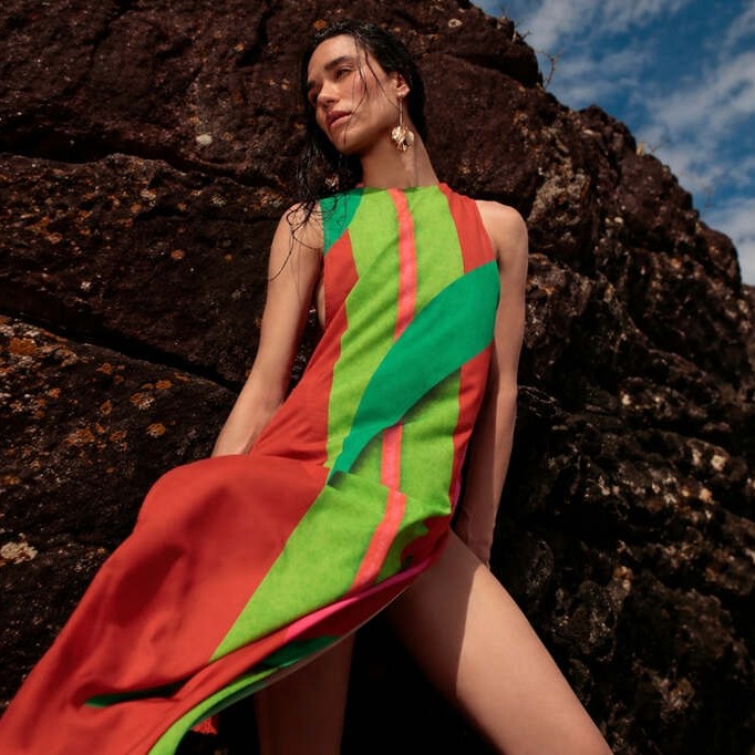 Mulher usando vestido leve estampado De Chelles encostada em pedra e com céu azul ao fundo, representando como usar saída de praia
