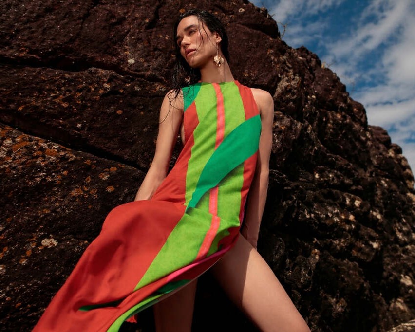 Mulher usando vestido leve estampado De Chelles encostada em pedra e com céu azul ao fundo, representando como usar saída de praia