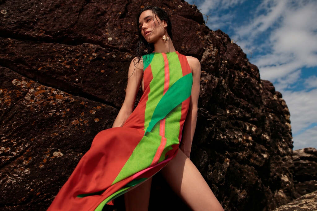 Mulher usando vestido leve estampado De Chelles encostada em pedra e com céu azul ao fundo, representando como usar saída de praia