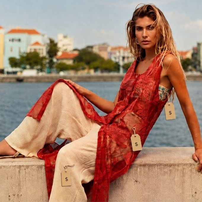 Modelo sentada em um muro pequeno com o mar e com paisagem ao fundo usando peças De Chelles com etiquetas de preço, representando como precificar roupas para revenda