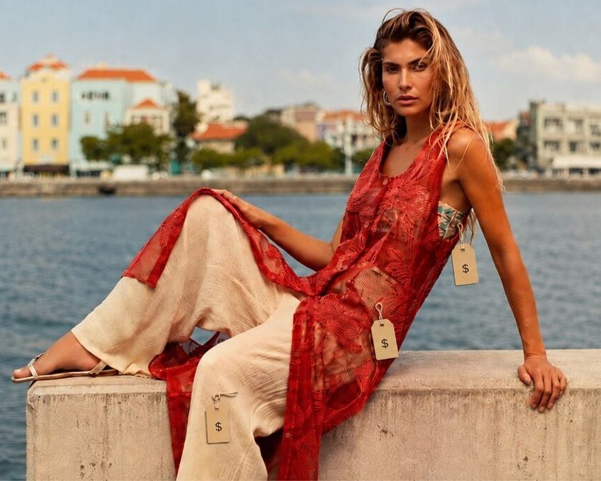 Modelo sentada em um muro pequeno com o mar e com paisagem ao fundo usando peças De Chelles com etiquetas de preço, representando como precificar roupas para revenda
